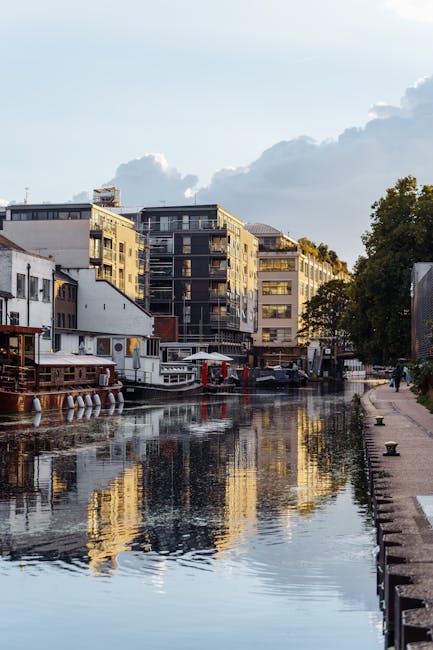 A canal scene featuring several narrowboats moored along the water's edge, with some partially obscured by trees with autumn foliage in shades of green, orange, and red. The boats are made of metal wi