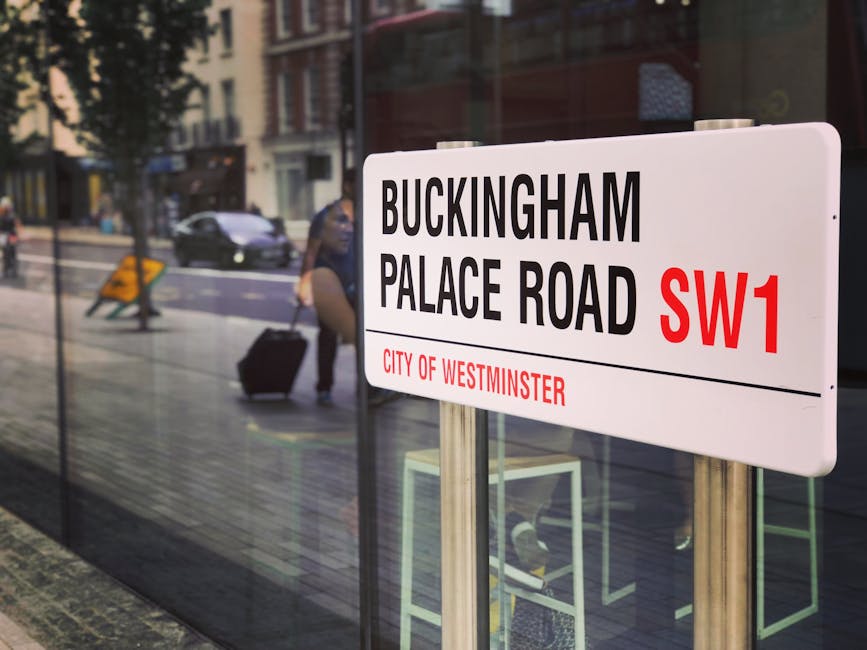A rectangular street sign mounted on metallic posts displays the location 'Buckinghampalace Road SW1' with black lettering on a white background, and the abbreviation 'SW1' in bold red font. The lower