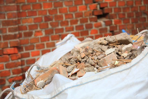 A large, white construction debris bag filled with broken bricks, shattered concrete, and wood fragments is positioned outdoors on a paved surface, with a brick wall in the background. The bag appears to be made of durable woven fabric, showing creases and slight dirt marks from use. The debris includes rough, irregularly shaped bricks and concrete pieces with varied shades of red, brown, and gray, some with visible surface textures like ridges or chipped edges. Thin, wooden sticks or dowels are also mixed within the debris. The scene is illuminated by natural daylight, casting soft shadows on the surrounding area, representing an environment typical of waste removal work in Westminster, where heavy building materials are being cleared. This image is relevant to rubbish removal services provided by Business Waste Removal Westminster, especially for construction or renovation waste disposal.